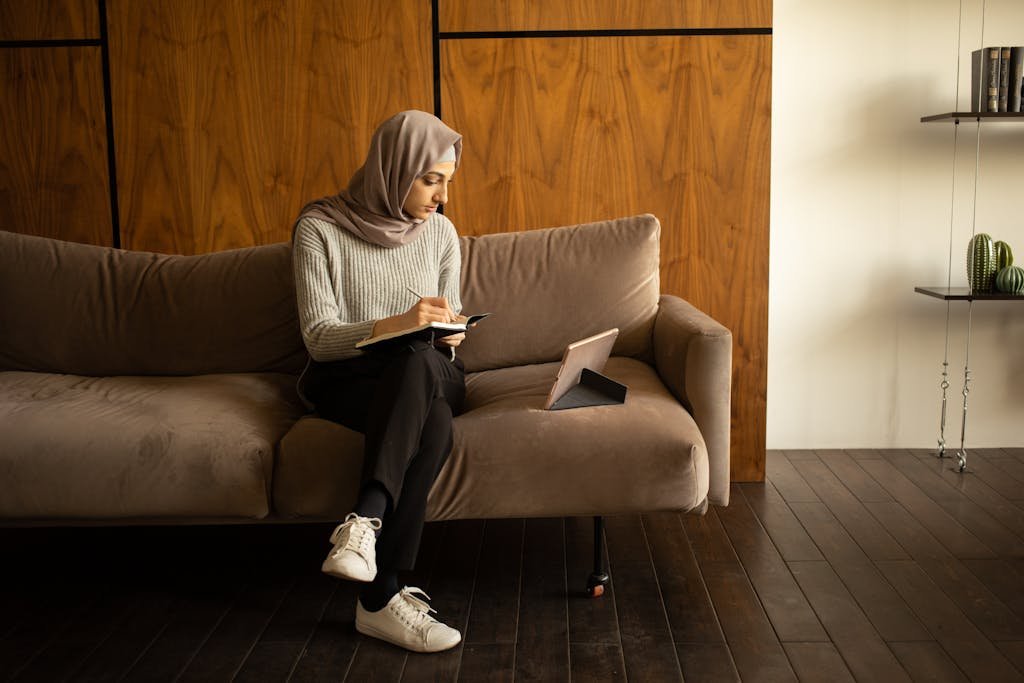 Focused Muslim woman writing in diary and watching tablet